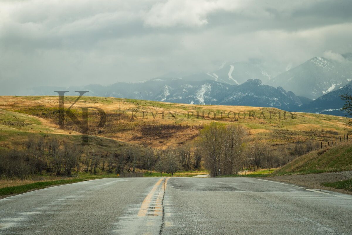 An open road leading toward snow-lined Montana peaks beneath dramatic clouds.