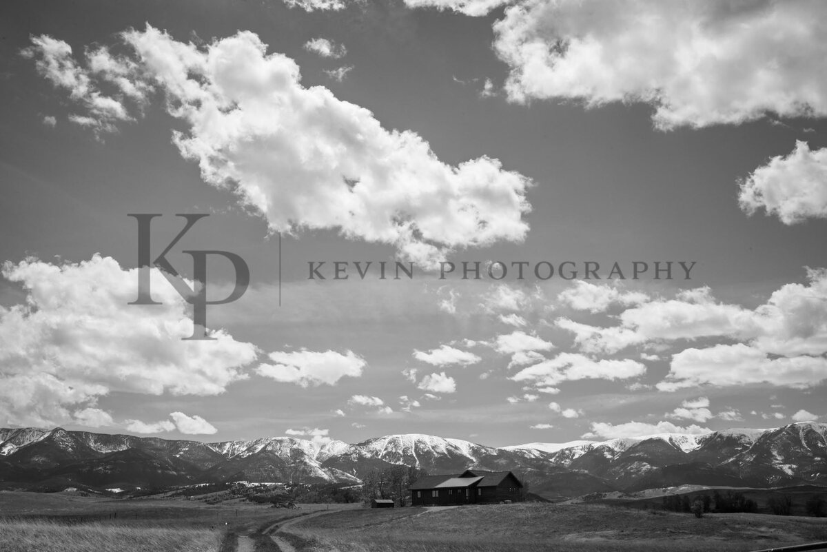 A monochrome homestead with rolling land and dramatic mountain clouds.
